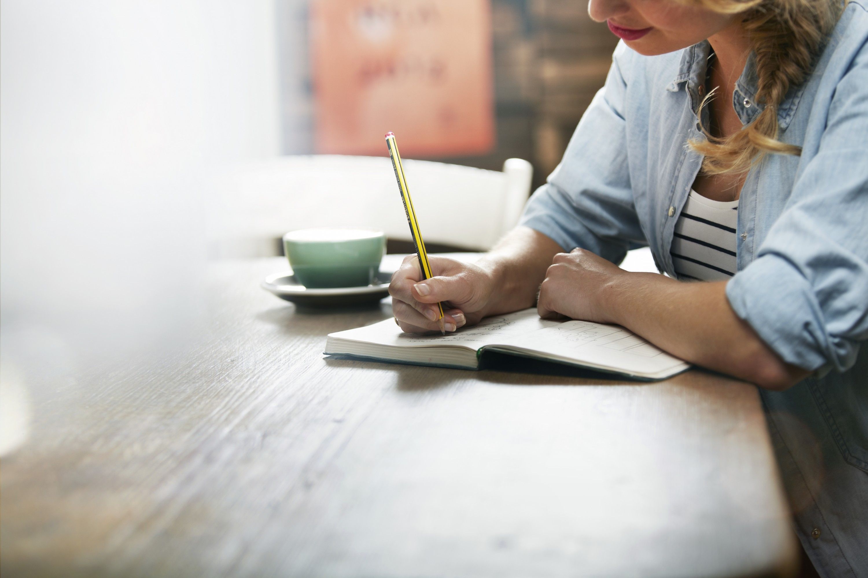 Uma mulher branca, loira, com uma trança escama de peixe ao lado do pescoço; está sentada em uma mesa de madeira, escrevendo em caderno com um lápis na cor dourada; ao lado do caderno tem uma xícara em cima de uma pires, nas cores verde- água. Uma mulher branca, loira, com uma trança escama de peixe ao lado do pescoço; está sentada em uma mesa de madeira, escrevendo em caderno com um lápis na cor dourada; ao lado do caderno tem uma xícara em cima de uma pires, nas cores verde- água.
