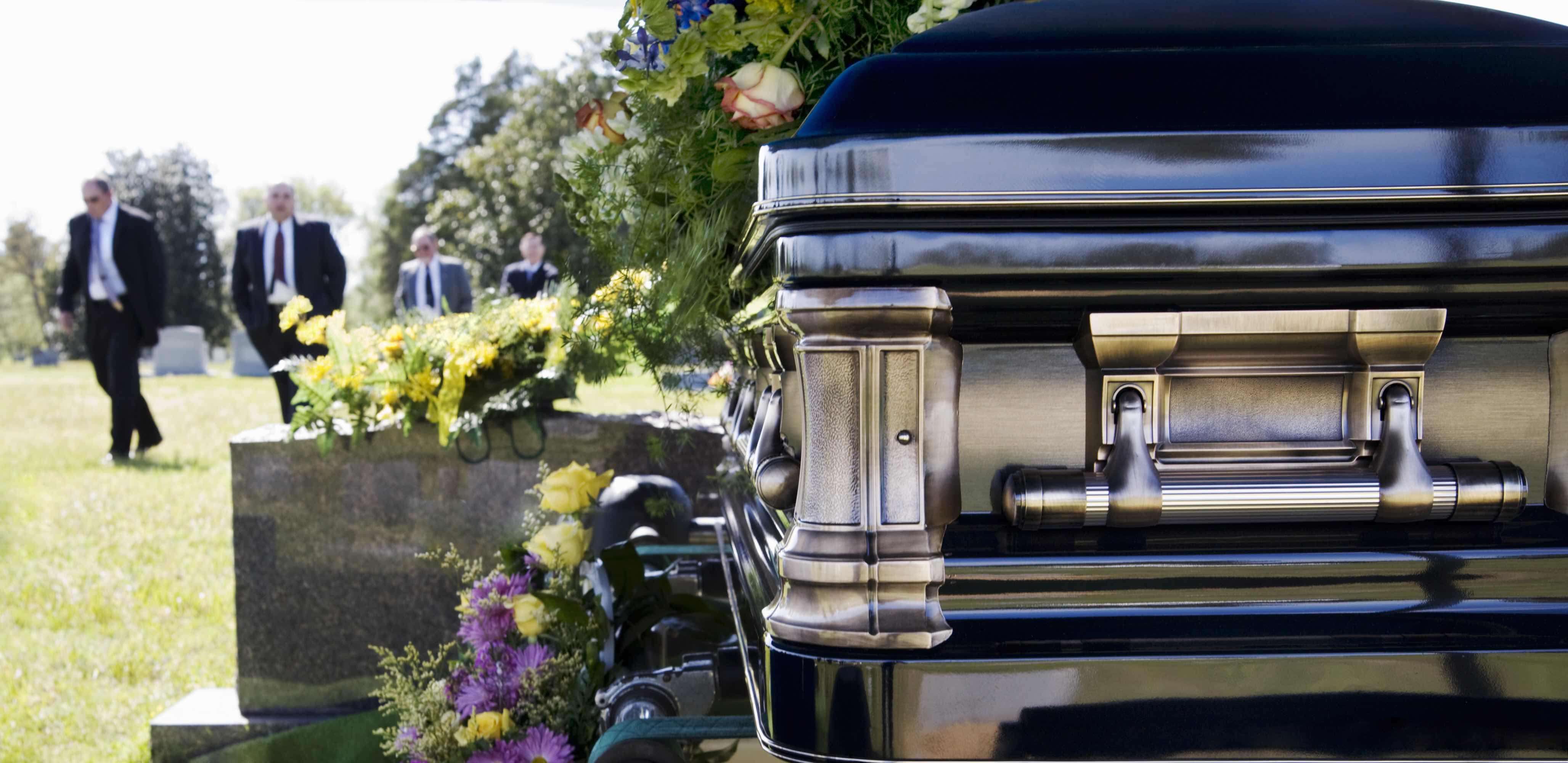 Nesta imagem em como organizar um funeral, temos em primeiro plano um caixão na cor preta e detalhes acobreados, coberto por flores. No fundo da imagem 4 pessoas (uma atrás da outra) vestindo terno. Nesta imagem em como organizar um funeral, temos em primeiro plano um caixão na cor preta e detalhes acobreados, coberto por flores. No fundo da imagem 4 pessoas (uma atrás da outra) vestindo terno.