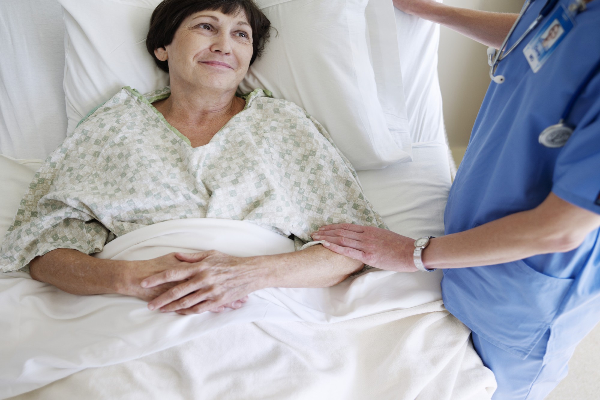 Uma senhora de cabelo curto castanho, está deitada em uma cama de hospital; ao seu lado temos uma enfermeira reconfortando.